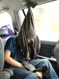Boy tries to sleep in car by hanging sweatshirt. 